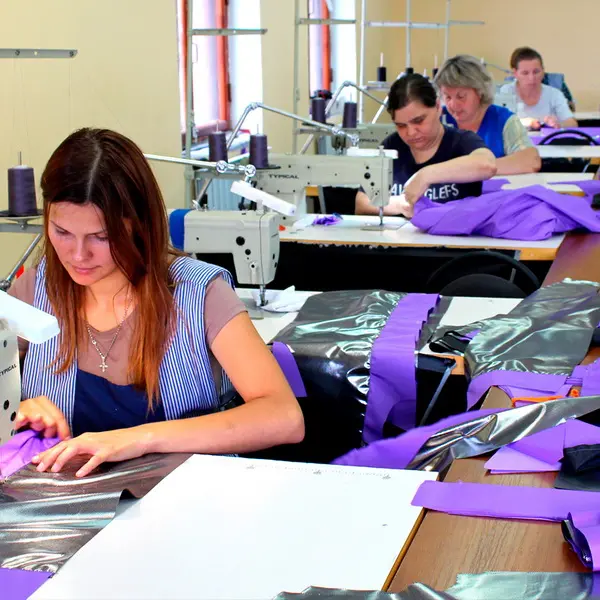 Женщины-швеи работают на промышленных швейных машинах, изготавливая партию спецодежды из фиолетовой ткани